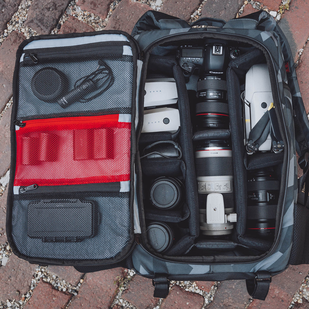 Glacier Cinema Backpack, Grey Camo. Front view with main compartment open. Photo shows camera accessories laid out in the bag.