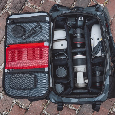 Glacier Cinema Backpack, Grey Camo. Front view with main compartment open. Photo shows camera accessories laid out in the bag.